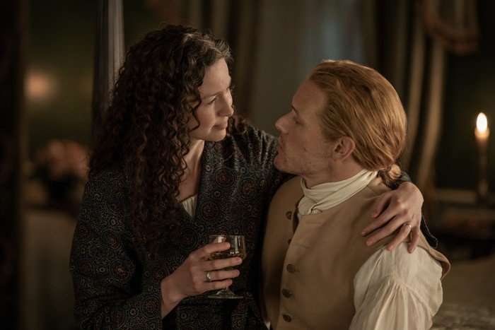 Caitriona Balfe as Claire Fraser and Sam Heughan as Jamie Fraser in Outlander season 7 part 2 in a close embrace while sat down in a dimly lit room.