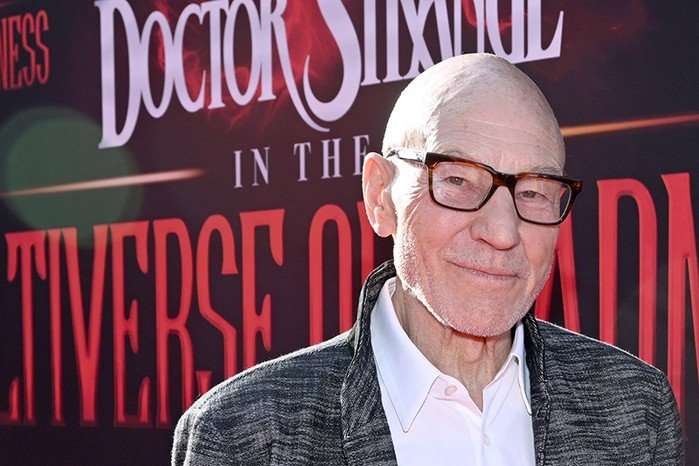 Sir Patrick Stewart attends the Doctor Strange in the Multiverse of Madness World Premiere, weating a shirt, jacket and glasses, standing in front of the film's logo