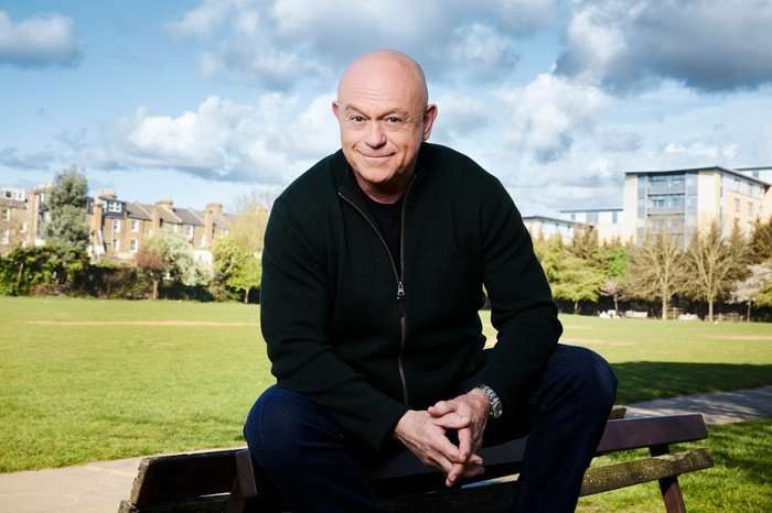 Ross Kemp dressed black sitting on the top of a bench in a park on a sunny but cloud-filled day