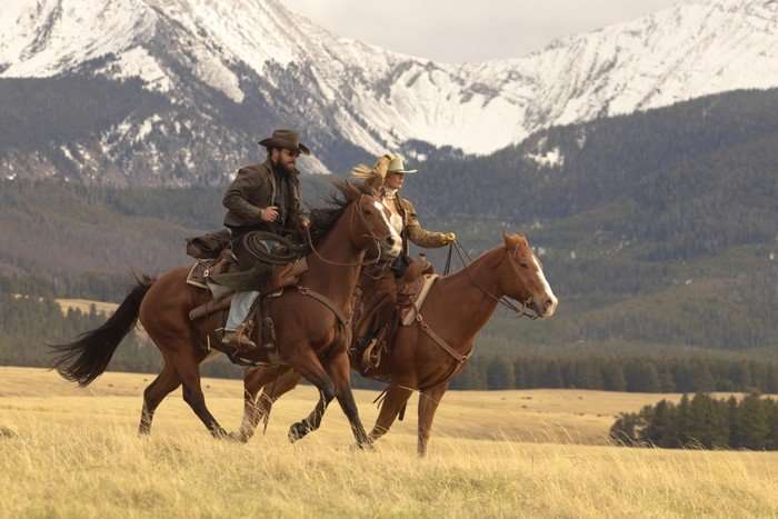 A man in a cowboy hat and a blond woman ride horses across a field. There are snow-covered mountains behind them
