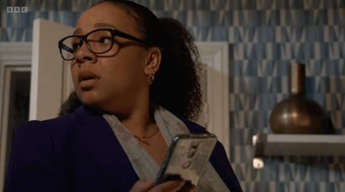 Belinda Owusu as Libby Fox holding a phone in EastEnders and looking behind her.
