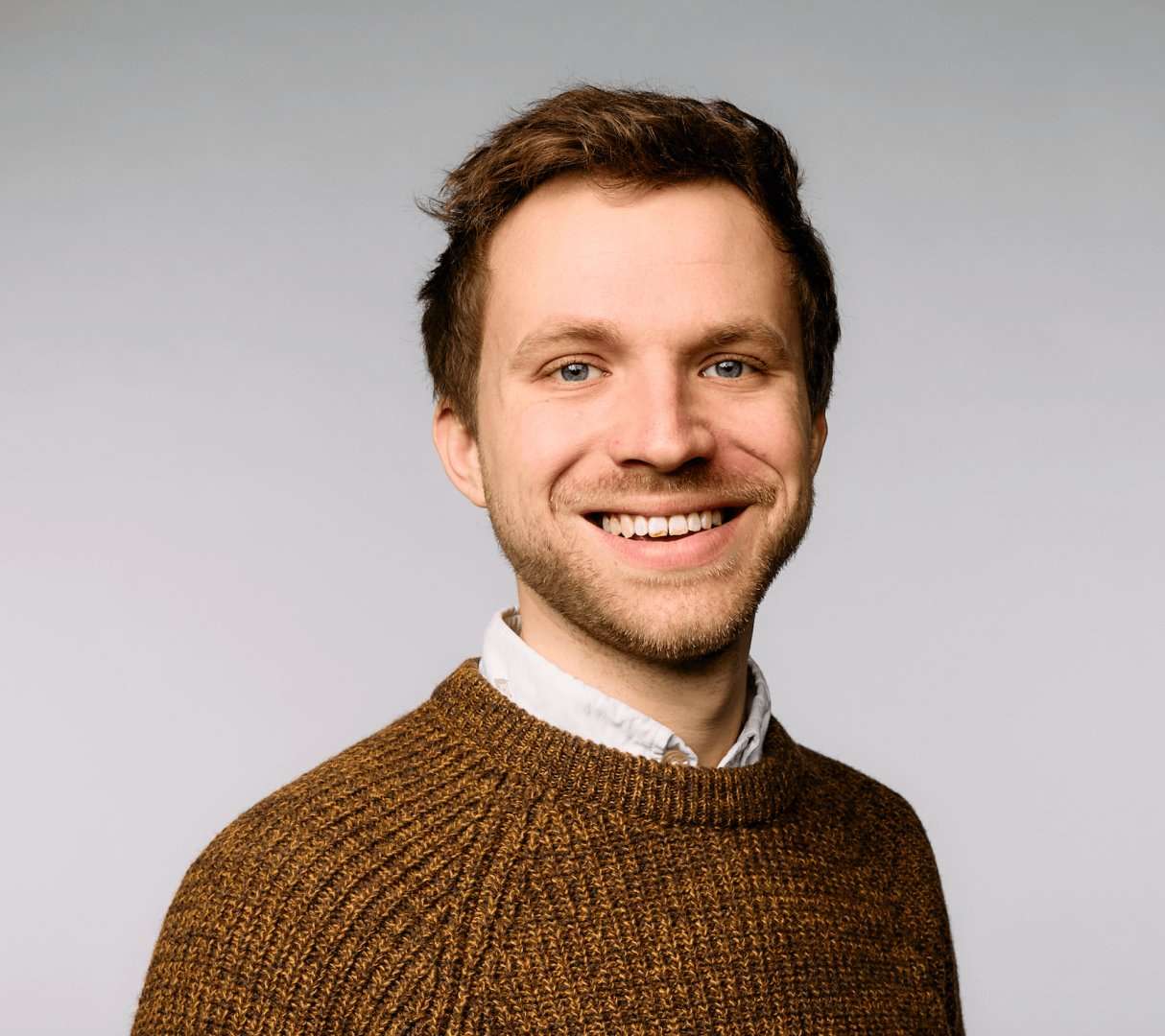 James Hibbs stands before a grey background, smiling and looking at the camera. He is wearing an orange-brown jumper over a white, buttoned shirt