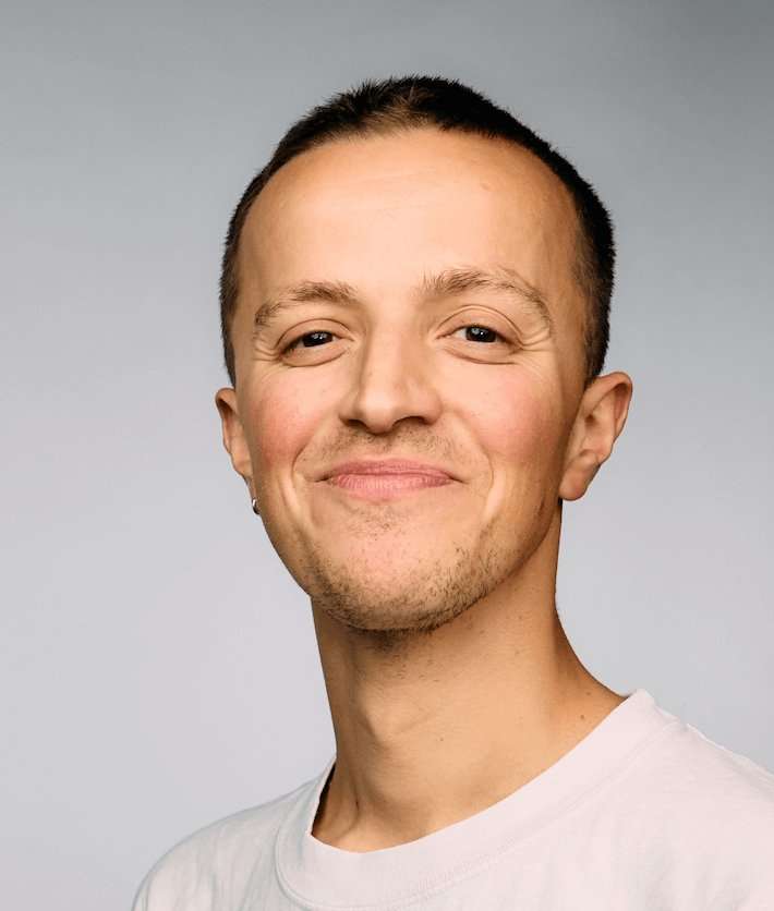 A head and shoulders shot of Ned Holmes. He is in front of a grey background, looking at the camera and smiling. He wears a white T-shirt