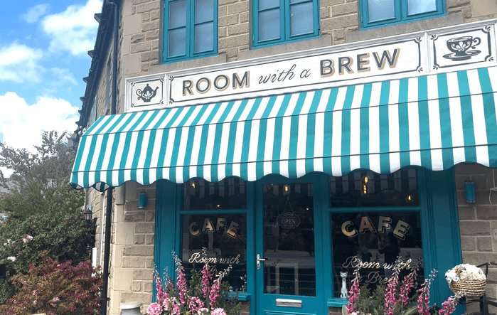 A shop front with white and turquoise coverings. A white sign above reads 'room with a brew' while gold painted signage on the window reads "cafe"