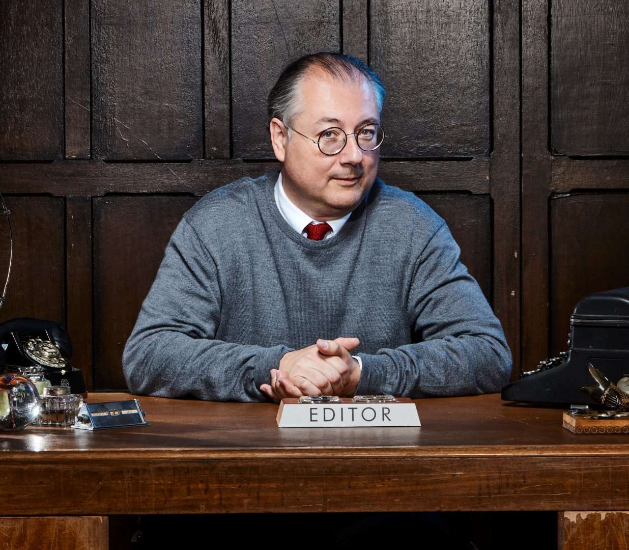Radio Times editor Shem Law wears a shirt, tie and grey jumper. He is sitting at a wooden desk behind a sign saying "Editor" clasping his hands. There are other items on the desk, including a dial telephone and typewriter.