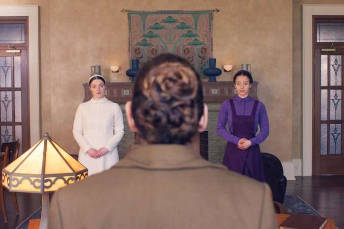 From behind a seated figure, two young women stand side by side in a formal room&mdash;one dressed in white, the other in purple&mdash;hands clasped as they face forward beneath a decorative wall hanging.