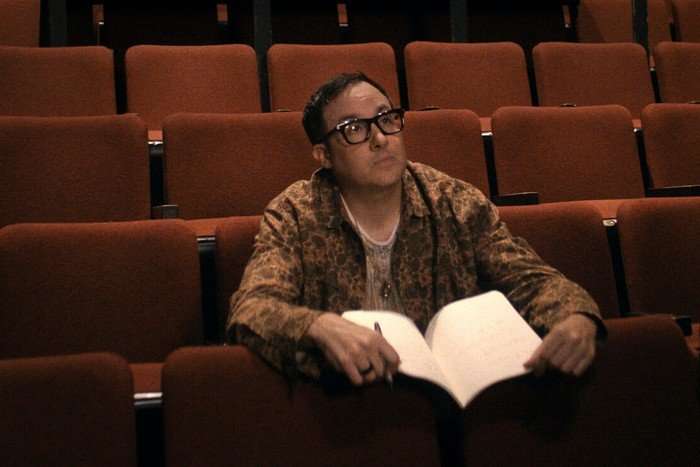 PJ Byrne as Adam in The Boys, sat in a row of red theatre chairs holding a script.