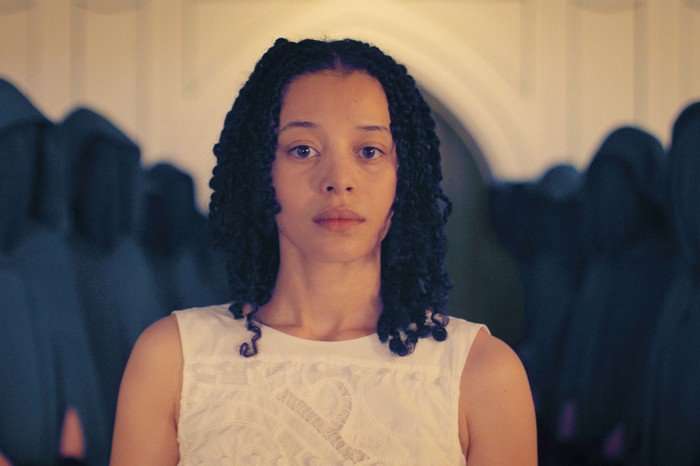 A woman with shoulder-length curly hair stands facing forward, wearing a white sleeveless dress, her expression calm and serious as blurred figures in dark clothing gather behind her.