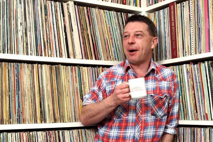 A main in a plaid shirt stands holding a mug and looks to the left in front of full shelves of vinyl records.