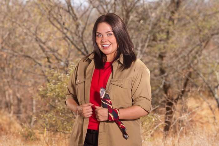 A woman stood in a safari landscape smiling ahead holding the lapels of her jacket.