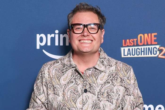 Alan Carr wearing a white and black patterned shirt, smiling widely ahead.