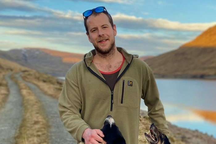 Alexander McKellar - a man wearing a green fleece and a black and red vest underneath - smiles in front of a picturesque countryside view.