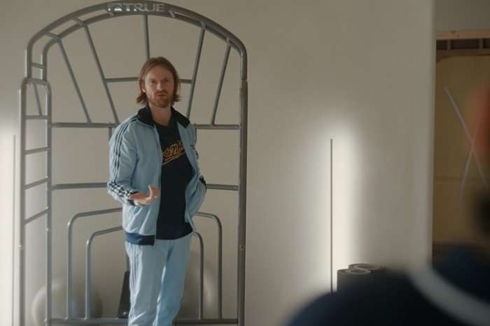 Finneas stands next to a workout frame wearing a tracksuit in a scene from Beef season 2