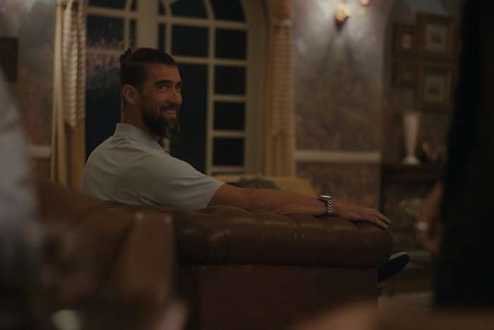 Michael Phelps sits on a sofa chair in a lounge, smiling at somebody behind him