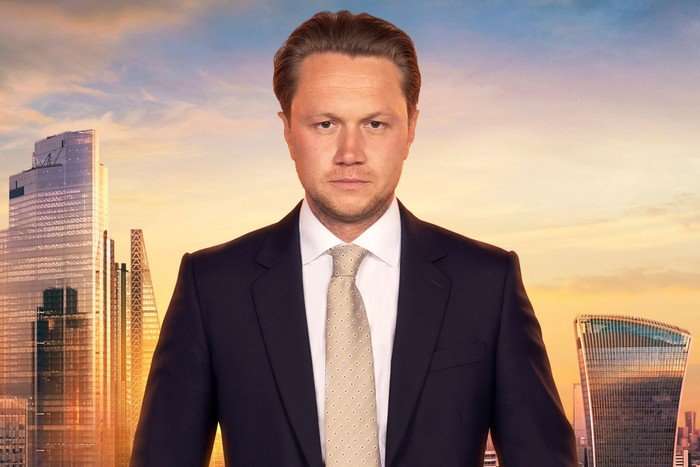 Dean wearing a black suit, looking seriously ahead.