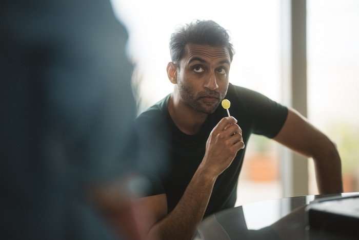 Avi Nash as Rav in Secret Service, sat down, holding a yellow lollipop.