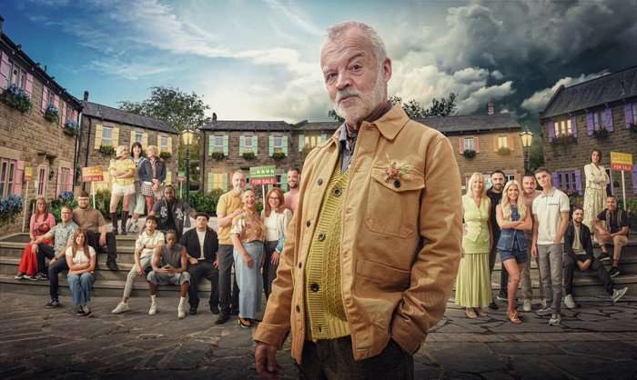 Graham Norton, in a tan jacket and jumper, stands in front of a group of people in a town square