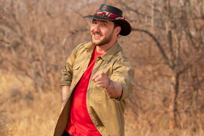 A man smiles ahead whilst stood with one hand on his hips and the other pointing forwards to the camera.