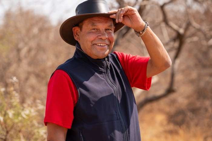 Craig Charles standing in a safari landscape and smiling ahead whilst holding the brim of his hat.