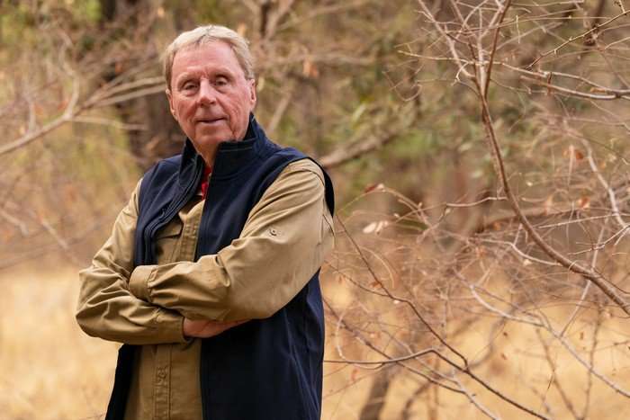 Harry Redknapp smiling ahead with his arms crossed in a safari landscape.