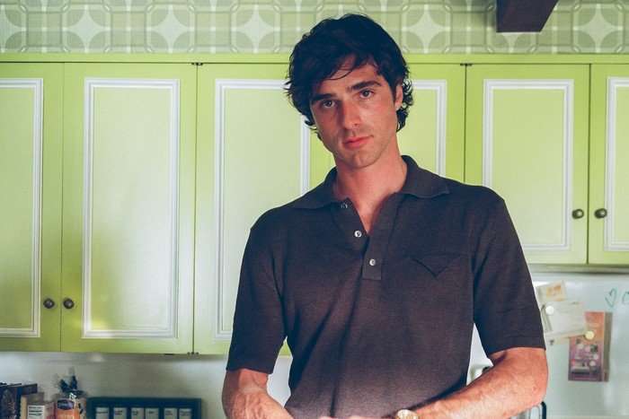Jacob Elordi standing in a kitchen with bright green cabinets, holding some food out of shot and wearing a brown polo shirt.