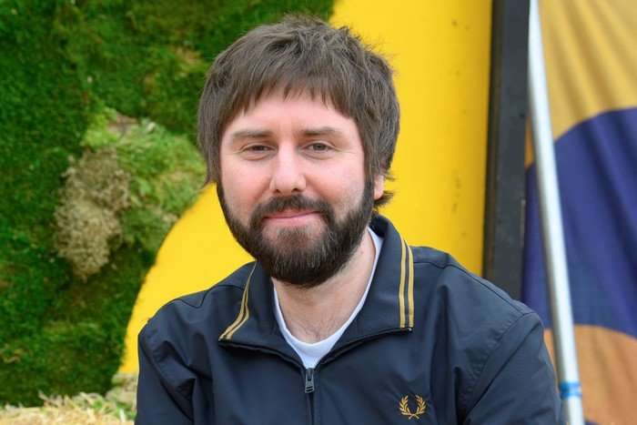 James Buckley smiling in front of a yellow and green background.