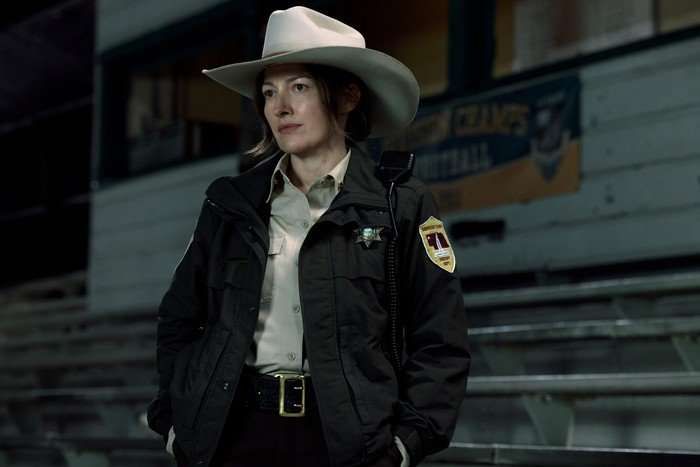 A sheriff wears her uniform and stands in a sports hall, looking sternly ahead
