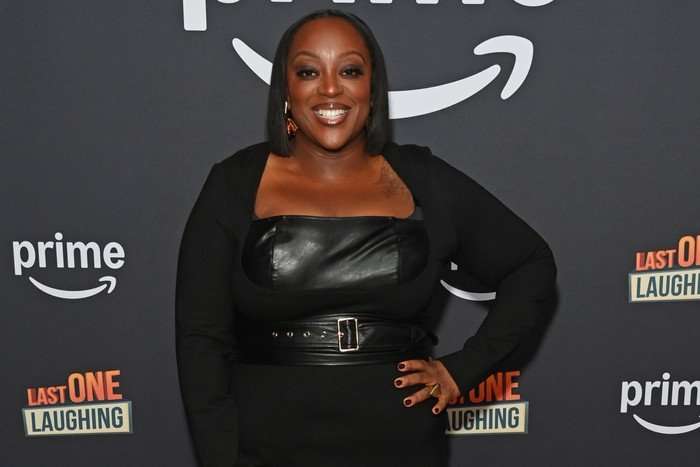 Judi Love wearing an all-black dress and is smiling while resting one hand on her hip.