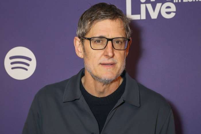 Louis Theroux wearing a black t-shirt and grey jacket. He is stood looking ahead in front of a purple background.