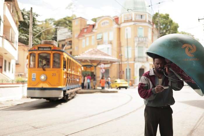 A man stands at a green public phone booth on a sunny street, holding a phone to his ear and looking down at a card in his hand. Behind him, a bright yellow tram approaches along curved tracks, with pastel-colored buildings and a small group of people gathered near a stop in the background.