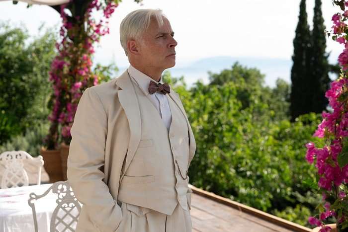 Mark Bonnar as Elmer Waysmith in Marble Hall Murders, standing next to a white table and looking out over trees, with bright plants either side of him.