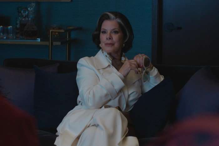 marcia-gay-harden-margo-money-troubles Elizabeth wears a white outfit and leans back on a sofa, looking bemusedly ahead of her and holding a pair of sunglasses