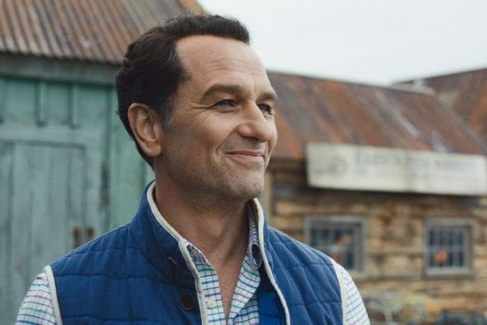 Matthew Rhys as Mayor Tom Loftis in Widow's Bay &ndash; he is wearing a shirt and gilet and smiles. He is standing in front of what appear to be old buildings with corrugated roofs
