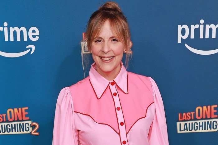 Mel Giedroyc smiling ahead as she poses in front of a dark blue background. She is wearing a two tone pink shirt and has her her hair styled in a bun.