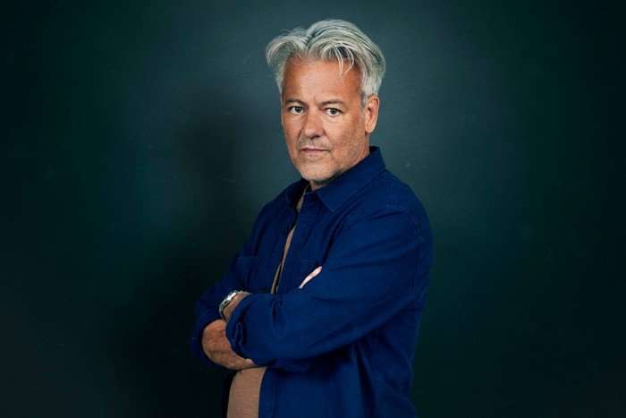 Rupert Graves as Jason Bradley in Missed Call, wearing a navy shirt and crossing his arms while standing in front of a blue backdrop.