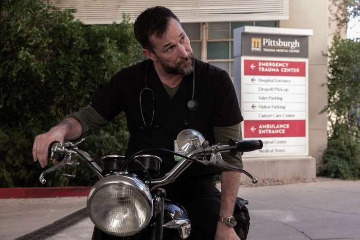 Noah Wyle as Dr Robby in The Pitt, sat on a motorbike while wearing his hospital scrubs.