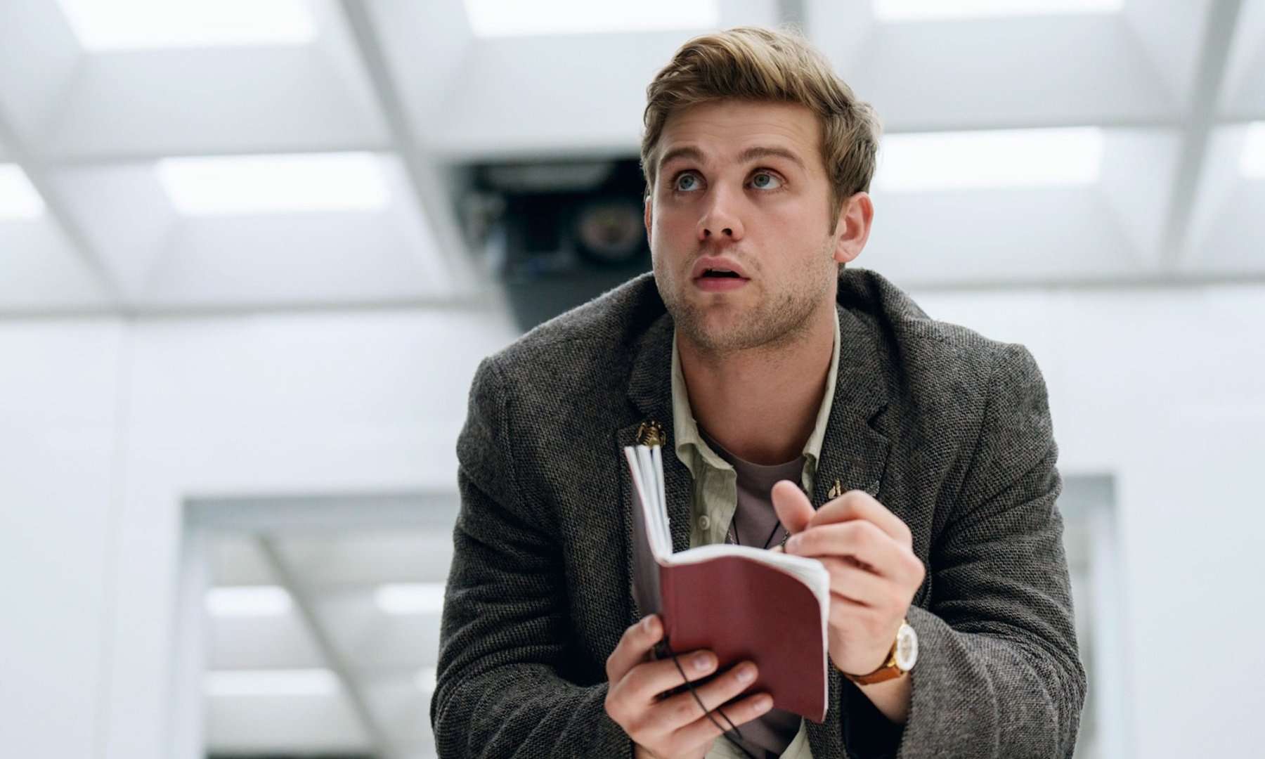 Leo Woodall as Edward sits in a white room with a notebook writing something down and looking up in Prime Target.