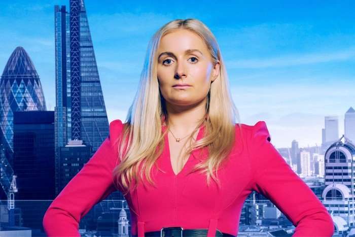 Rachel posing for The Apprentice 2024. She is wearing a pink dress and is in front of a background of the London skyline