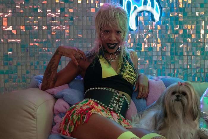 rico-nasty-margo-money-troubles Rico Nasty stars in Margo's Got Money Troubles; her character, KC, is wearing colourful clothing and leaning back on a sofa. The wall behind her has neon lights and shimmering blue tiles. A small dog is sat beside her.