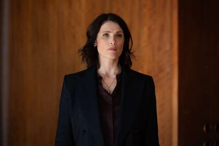 Gemma Arterton as Kate in Secret Service, wearing a black jacket and purple shirt, stood in front of a wooden wall.