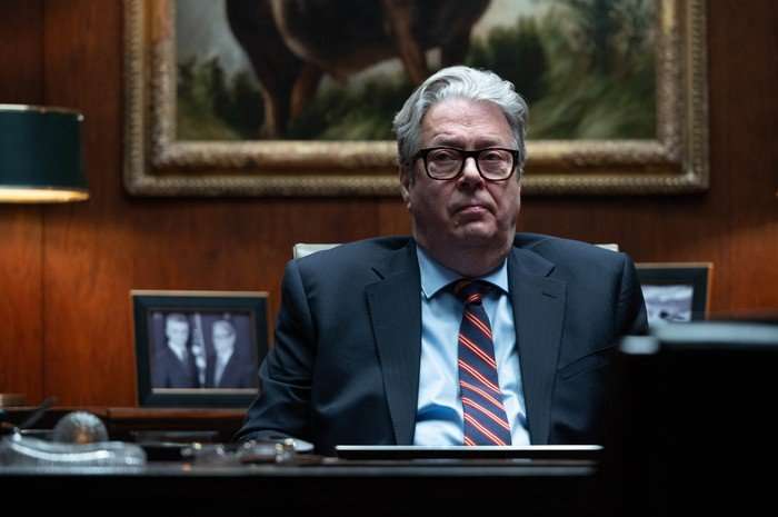 Roger Allam as Sir Alan in Secret Service, wearing a suit and sat behind a desk in an office.