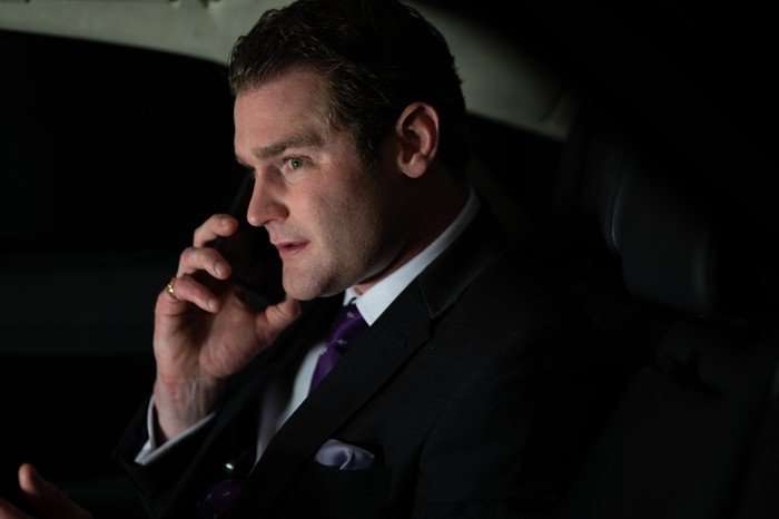 Mark Stanley as Ryan in Secret Service, sat in a car, wearing a suit and speaking on the phone.