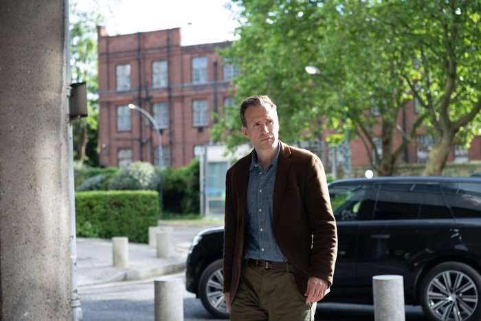 Rafe Spall as Stuart in Secret Service, walking outside with a car behind him.