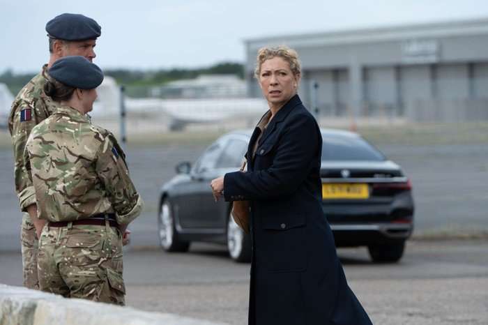 Alex Kingston as Rose in Secret Service, stood at an airport next to soldiers in uniforms, with a car parked behind her.