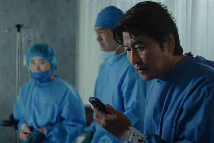 Song Kang-ho plays Dr Kim in Beef season 2; he is in an operating theatre wearing medical scrubs, with two assistants in the background behind him