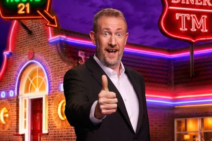 Alex Horne wearing a suit jacket and shirt, giving a thumbs-up sign and big smile, standing outside the Taskmaster house.
