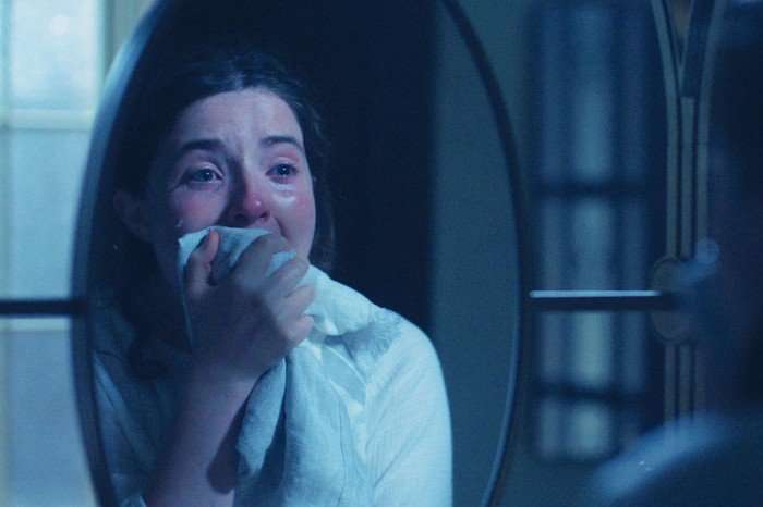 A tearful woman sits in a dimly lit room, holding a cloth to her mouth as she looks into a mirror, her eyes red and glistening with emotion.