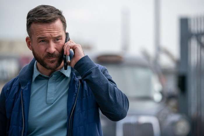 Barry Sloane answering a call and holding a phone up to his ear as he stands on the pavement with a taxi in the background.