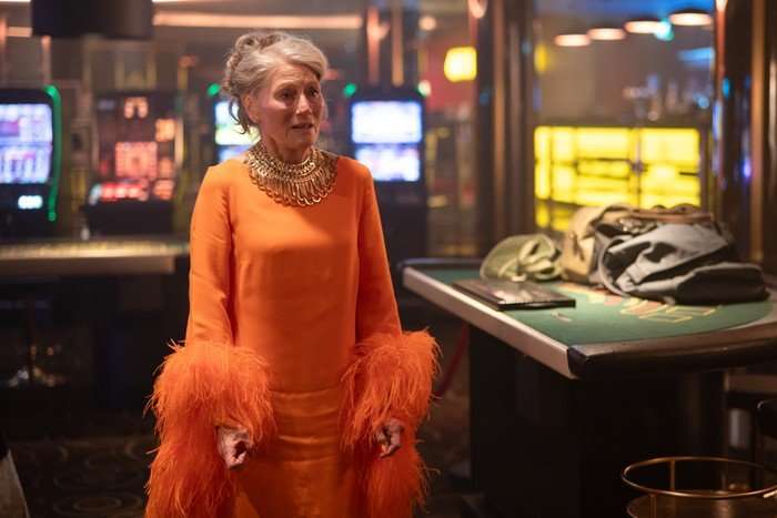 Geraldine James standing in the middle of a casino, wearing a bright orange dress with feather cuffs and jewellery, looking defeated and sad.