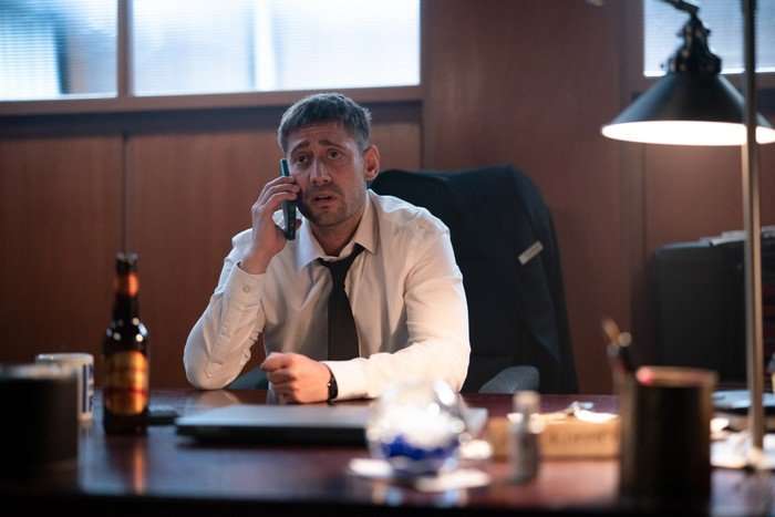 Michael Socha sitting at a desk, wearing a white shirt and tie looking stressed as he answers a call on the phone.